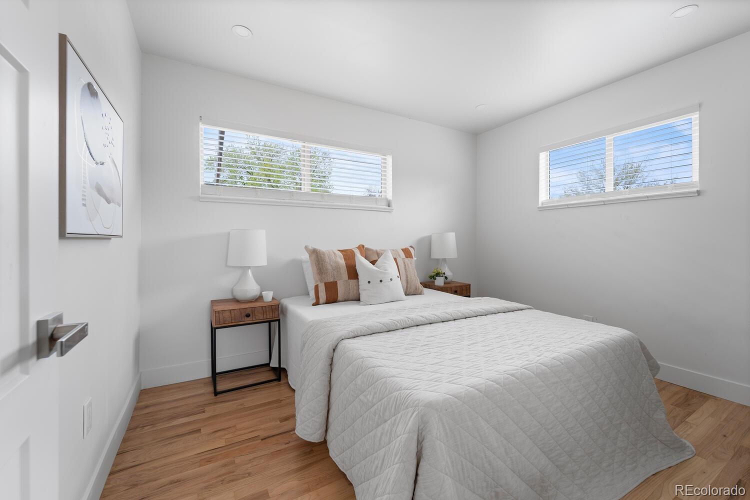 Bright bedroom with a white quilted bed, wood nightstands, and two high windows.