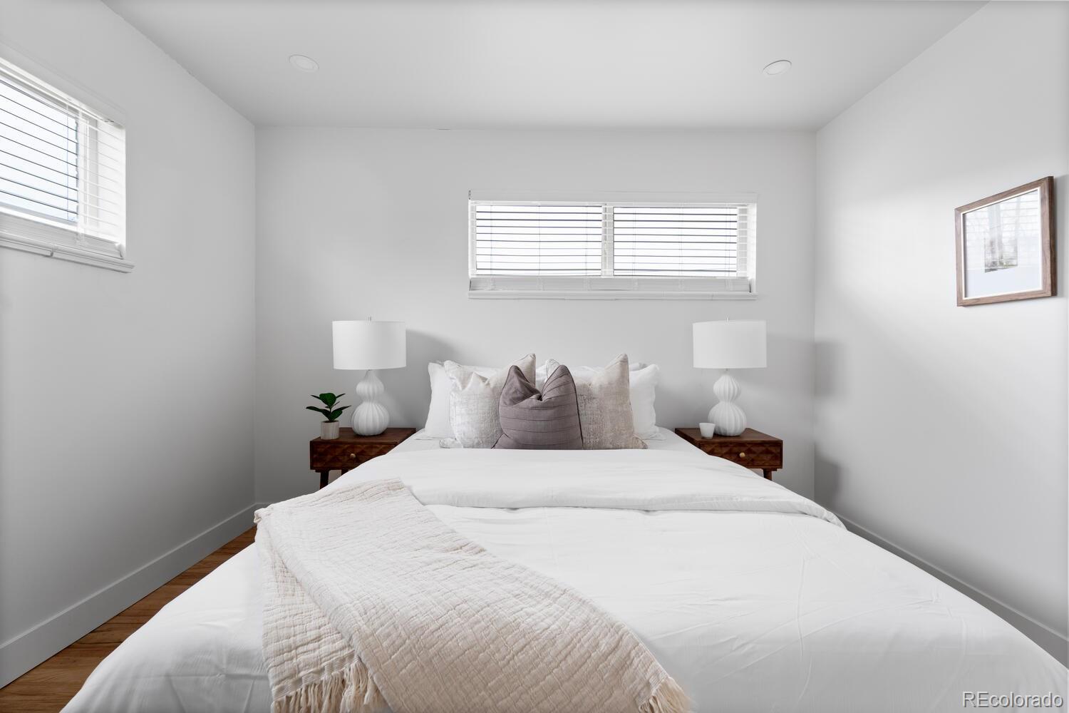 Staged bedroom with a white bed, neutral pillows, matching bedside tables, and windows with blinds.