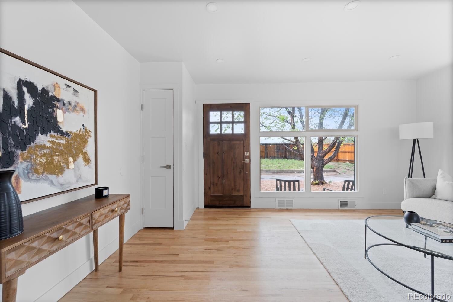 Staged living room with light hardwood floors, a dark wood front door, and a large window overlooking a tree.
