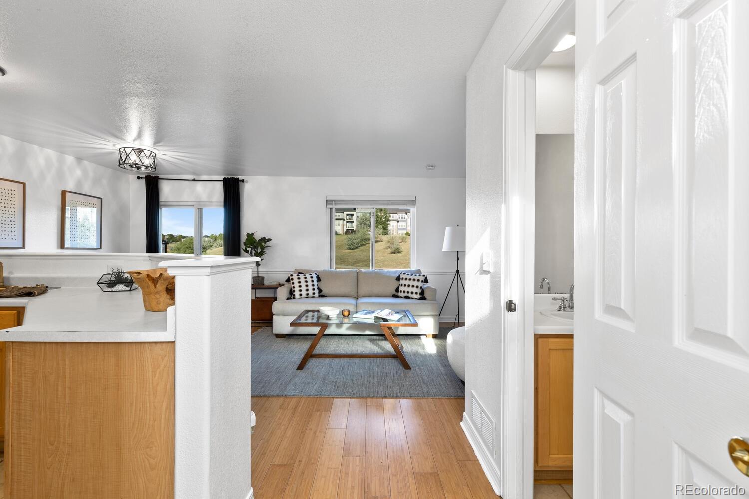 Staged living room with a light sofa, glass coffee table, large windows, and an adjoining kitchen counter.