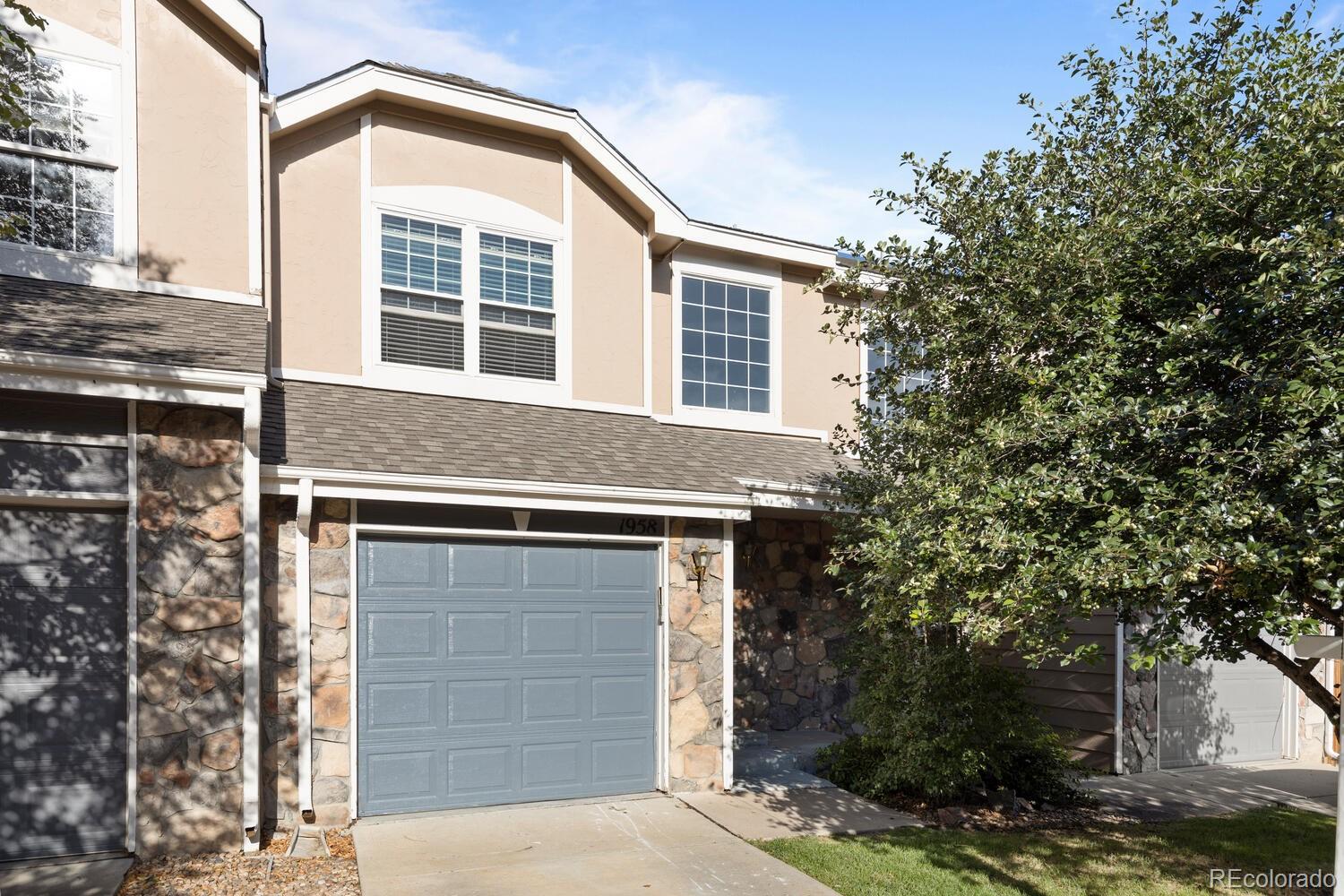 Two-story beige home with stone veneer, a blue-gray garage door, and leafy trees.