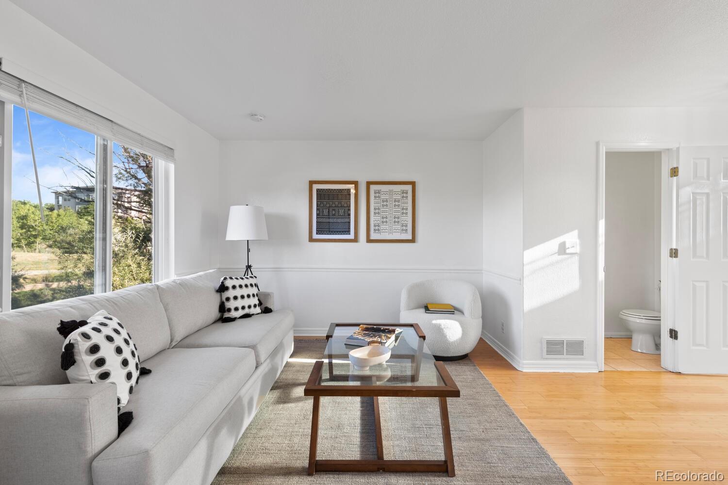 Bright living room with a gray sofa, glass coffee table, large windows, and a bathroom doorway.