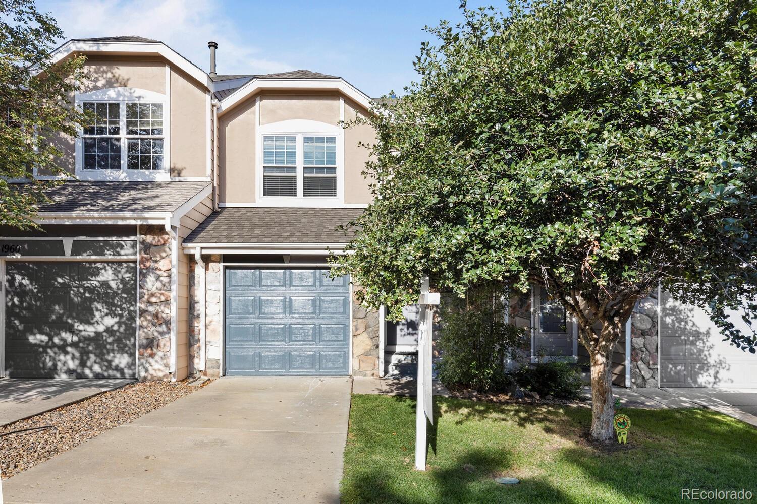 Two-story beige house with a blue-gray garage door, stone accents, and a mature tree in front.