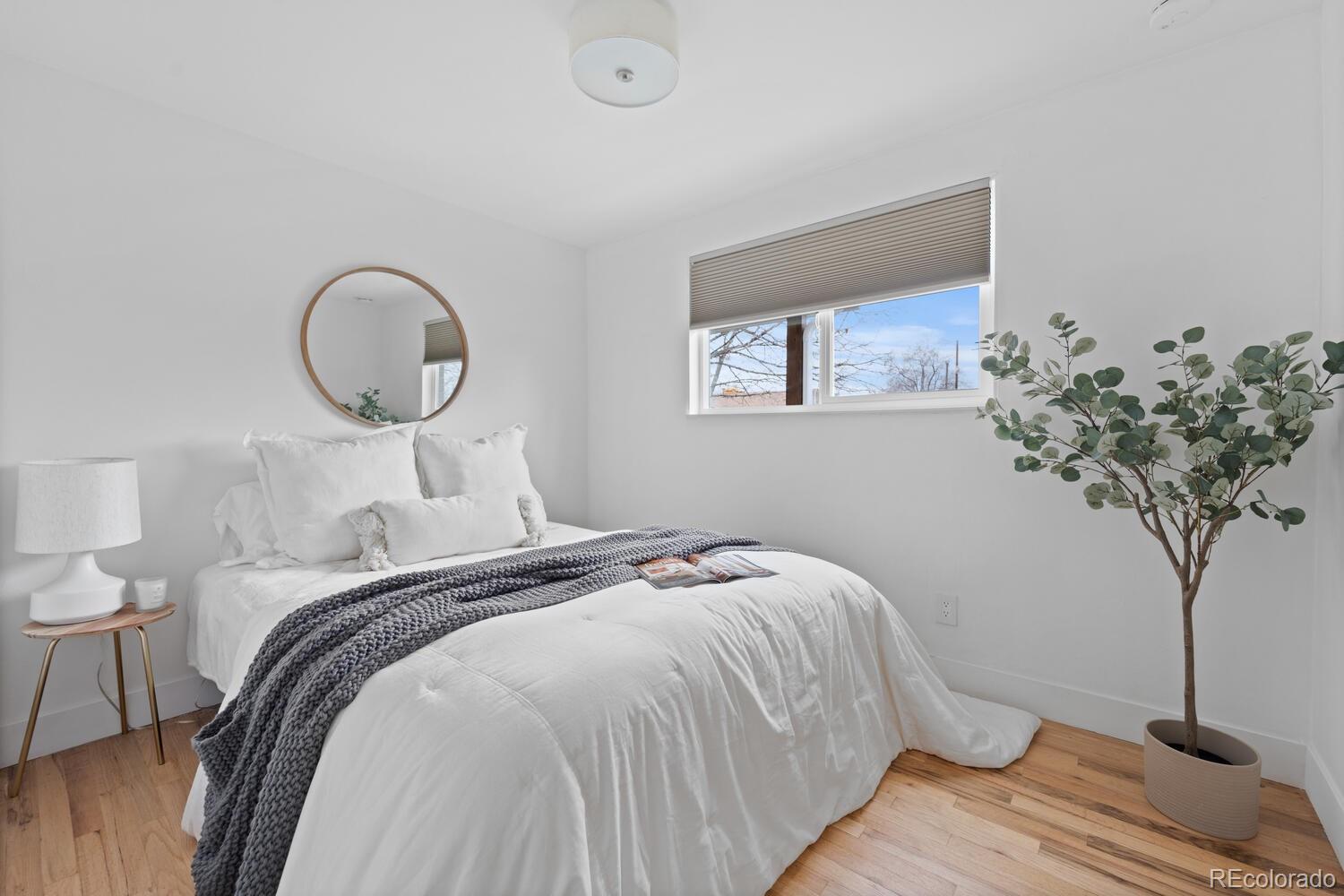 Staged bedroom with a white bed, charcoal knit throw, round mirror, and light wood floors.