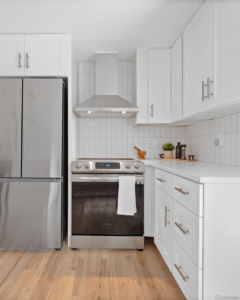 White kitchen with stainless steel refrigerator and range, tiled backsplash, and light wood flooring.