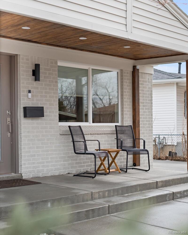 Front porch with two black chairs and a small table beneath a wood-paneled ceiling.