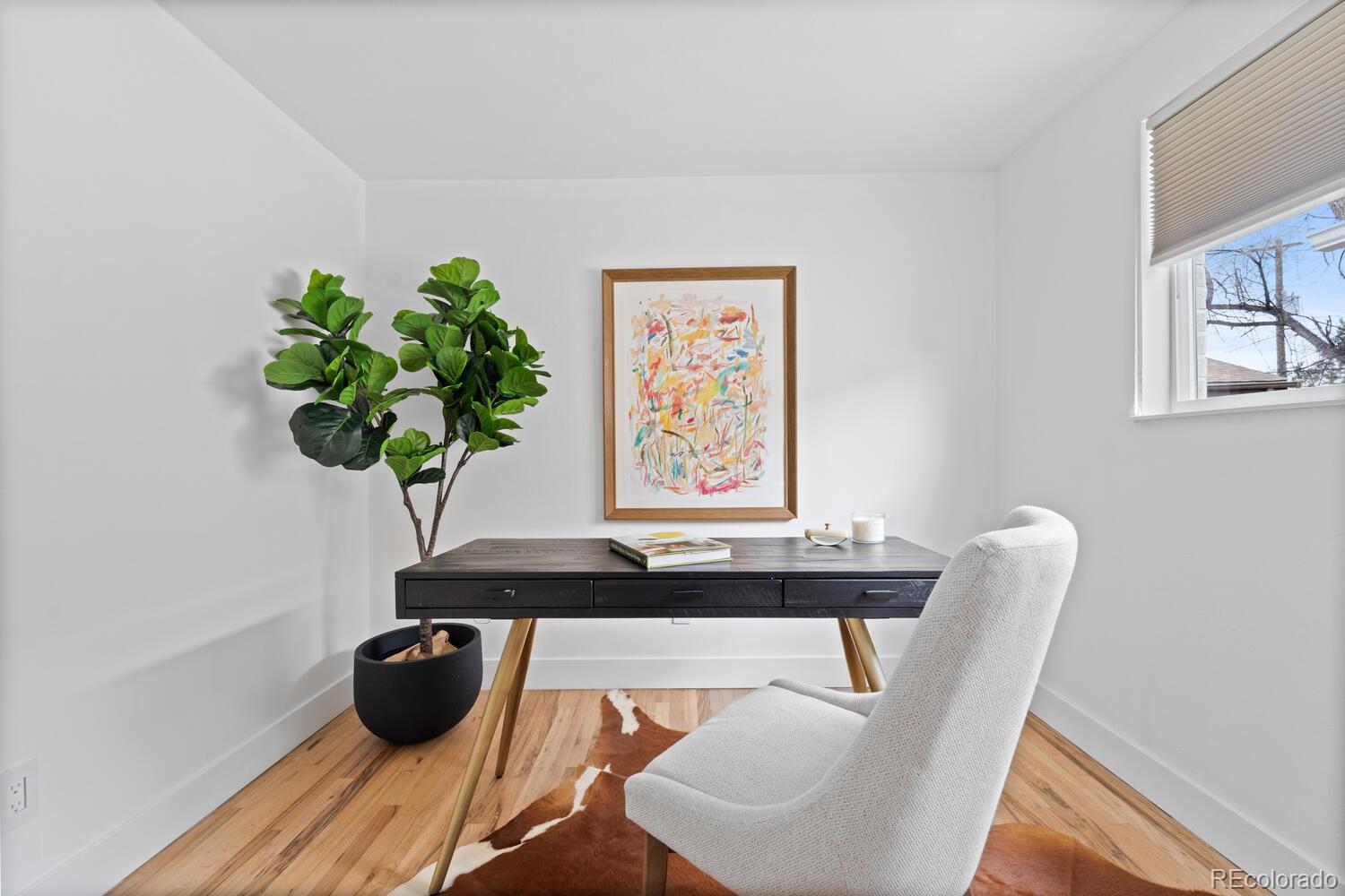 Furnished home office with a dark desk, white chair, potted plant, and framed abstract artwork.
