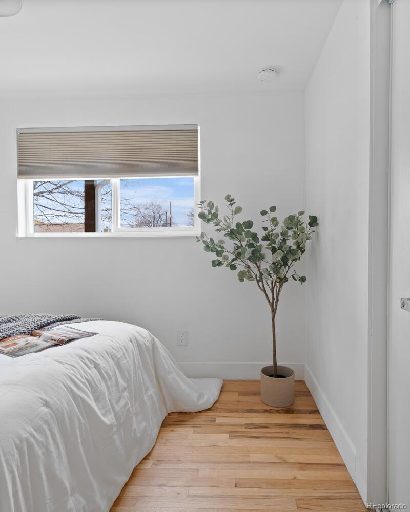 Bedroom with white bedding, hardwood floors, and a potted tree beside the window.