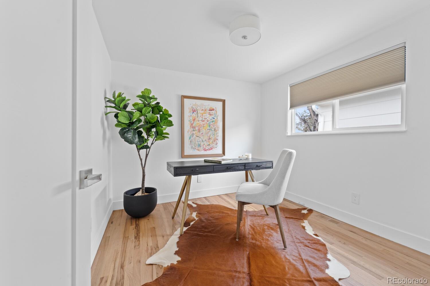 Staged home office with a dark desk, white chair, potted tree, and leather rug.