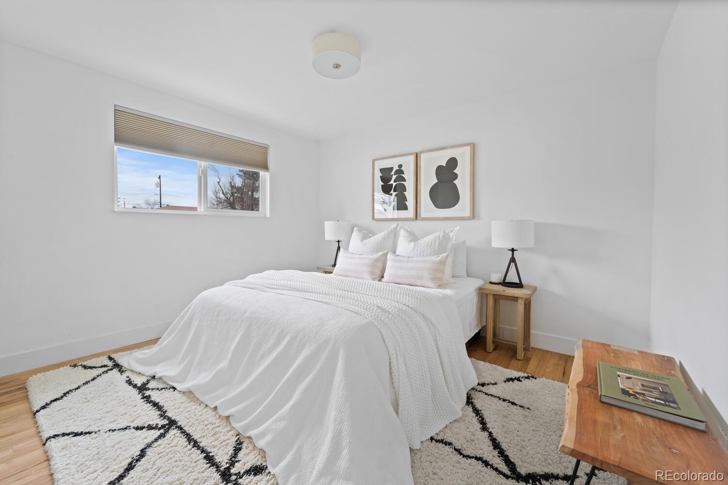 Staged bedroom with a white bed, geometric rug, abstract art, and a window with a pleated shade.