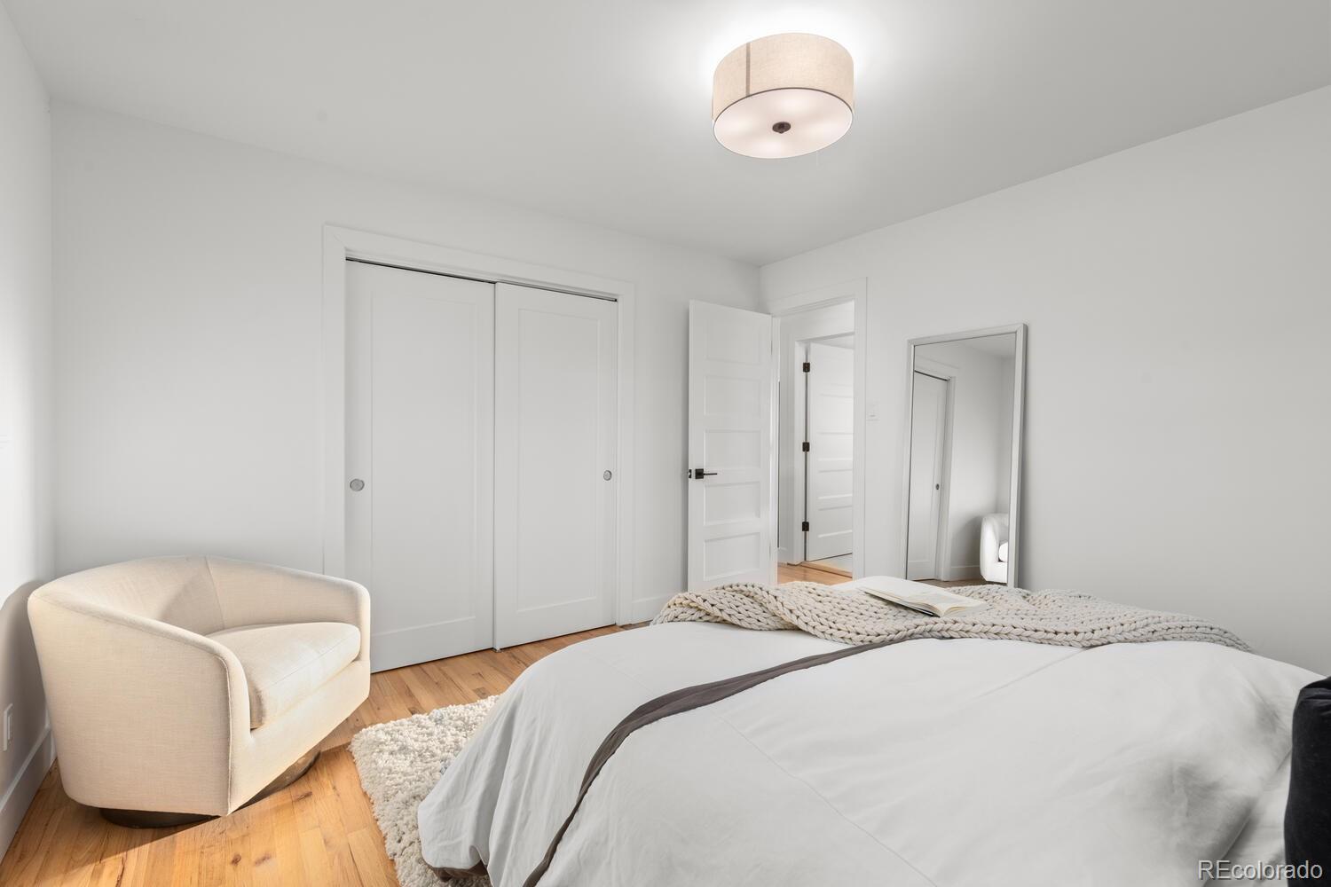 Staged bedroom with a white bed, cream armchair, hardwood floors, and sliding closet doors.