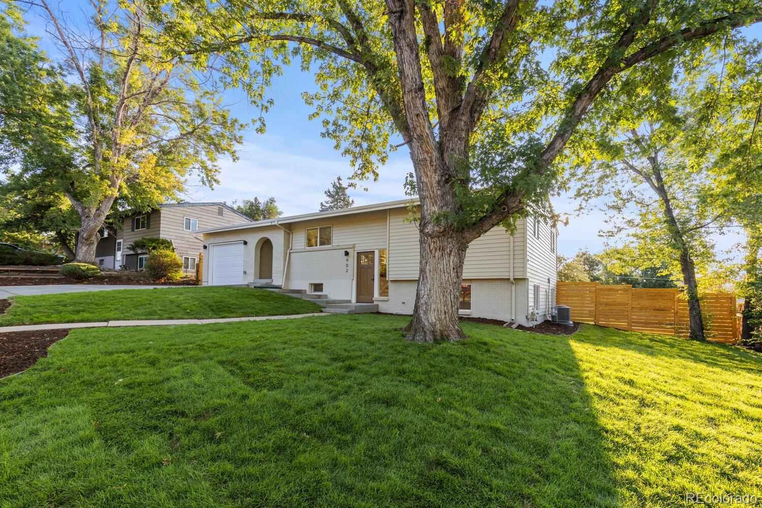 Home exterior with attached garage, broad lawn, mature shade trees, and wooden privacy fence.