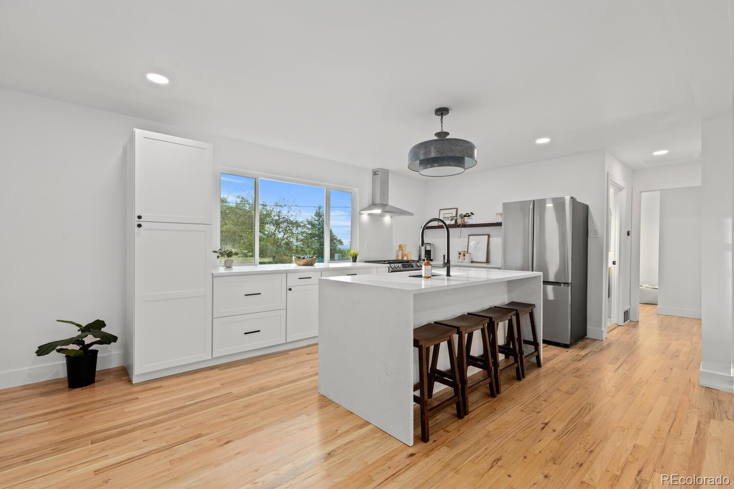 Bright white kitchen with a waterfall island, dark stools, stainless steel appliances, and hardwood floors.