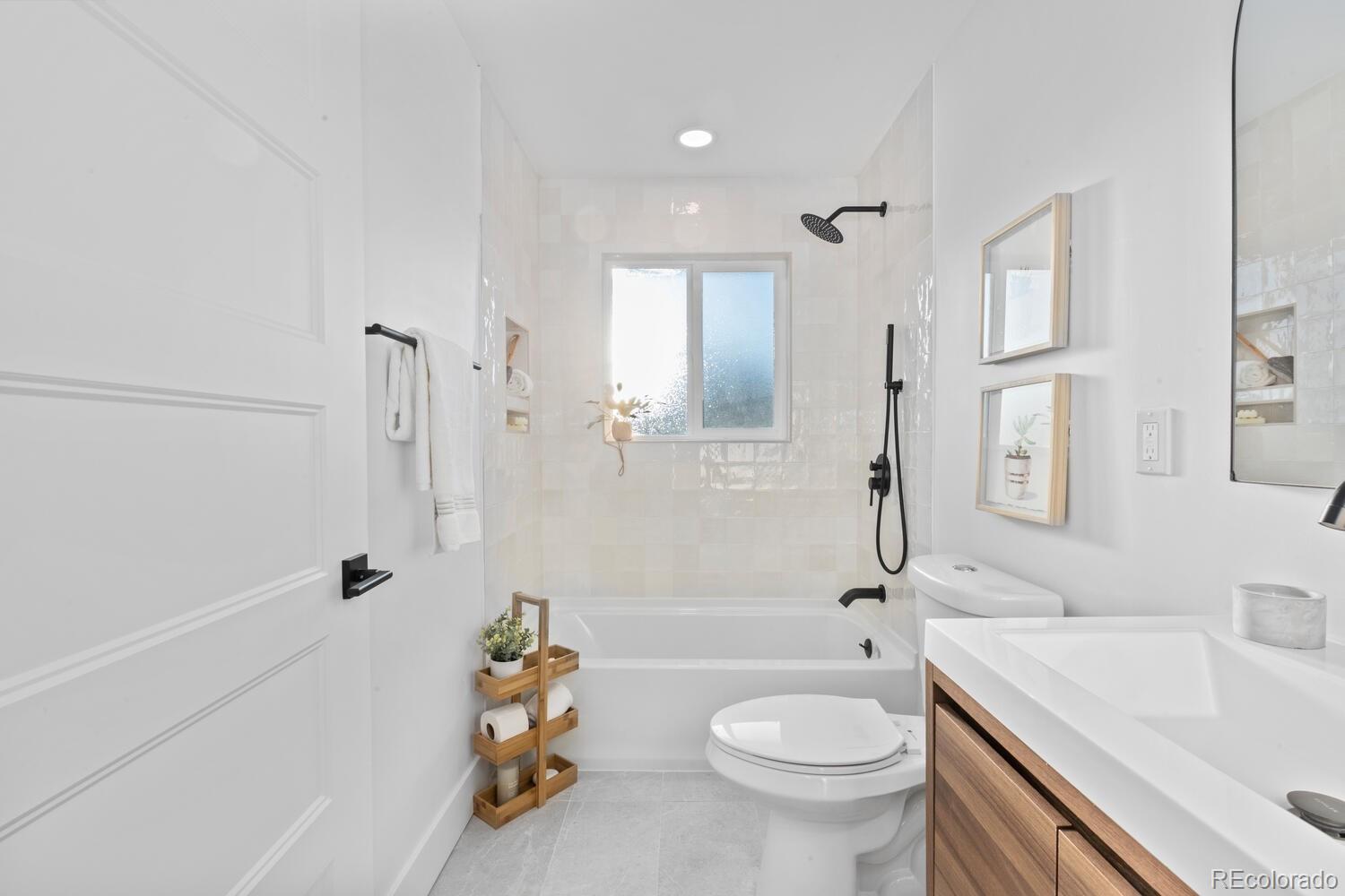 Bright bathroom with a tiled tub and shower, frosted window, black fixtures, and wood vanity.