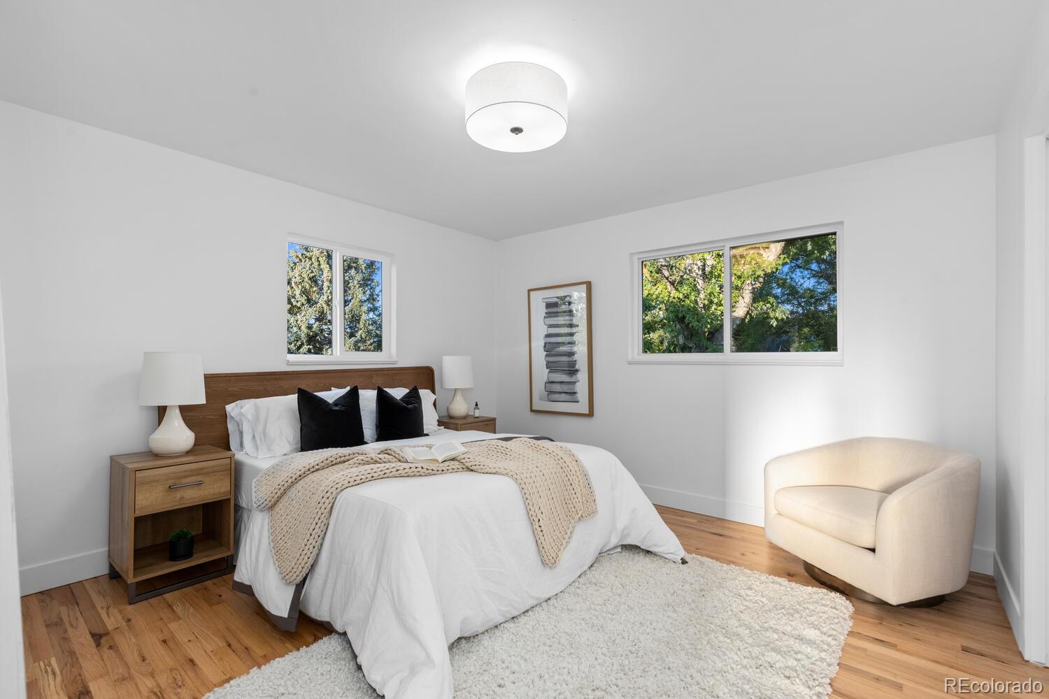 Staged bedroom with a white bed, black pillows, knit throw, and cream armchair beside two tree-facing windows.