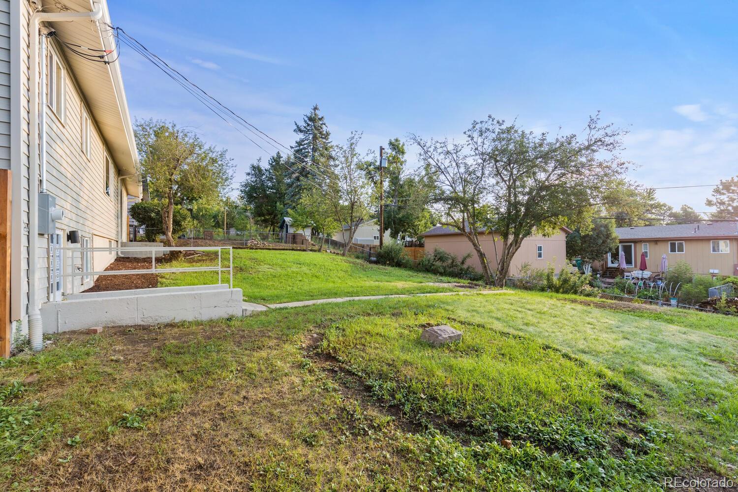Sloping backyard beside a light-colored house, with a retaining wall, railing, and mature trees.