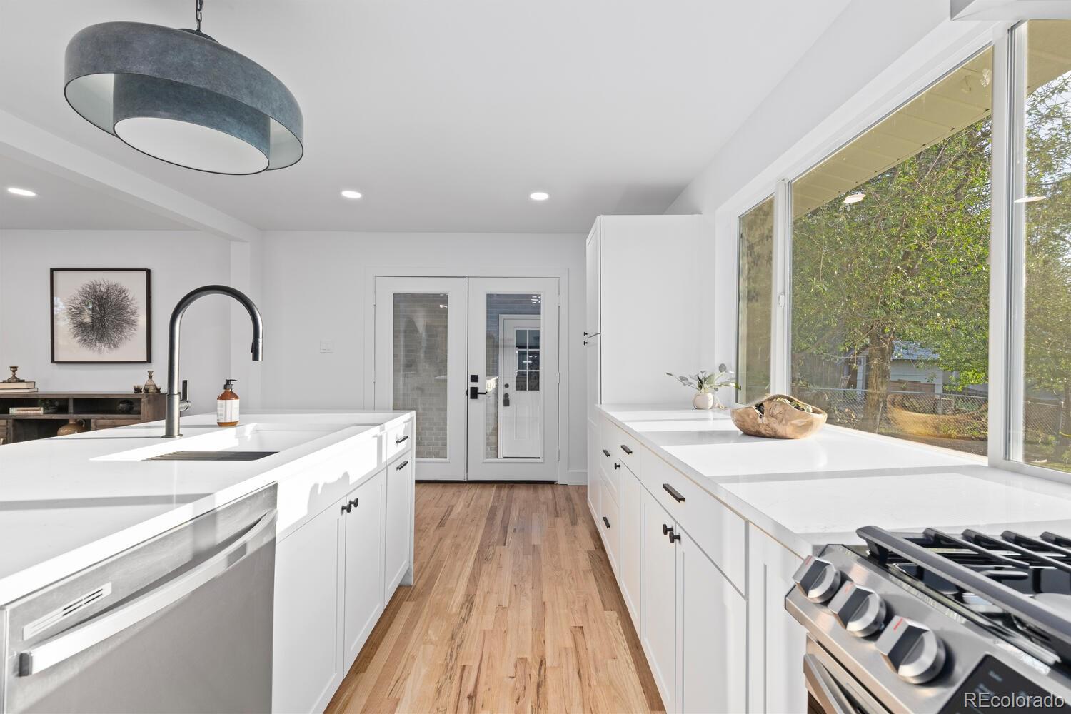 Bright white kitchen with stainless steel appliances, warm wood floors, and large windows overlooking trees.