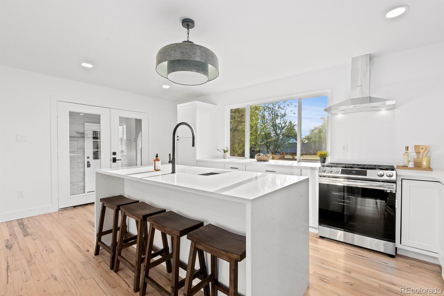 Modern white kitchen with a large island, dark wood stools, stainless-steel range, and wide garden-facing window.