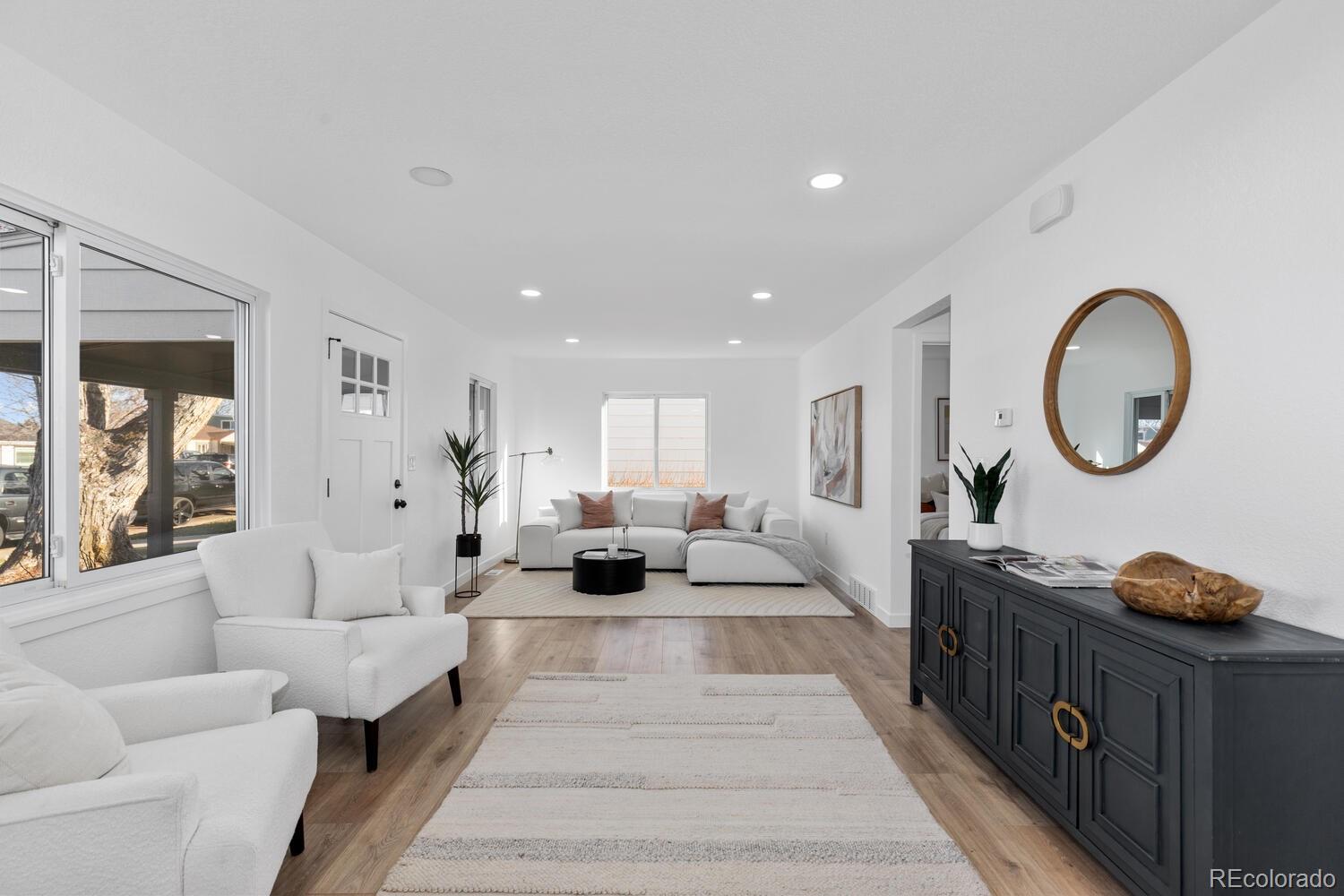 Staged living room with white sofas, light wood floors, and a dark console beneath a round mirror.