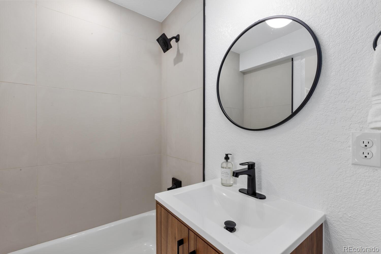 Bathroom vanity with a black faucet and round mirror beside a tiled bathtub.