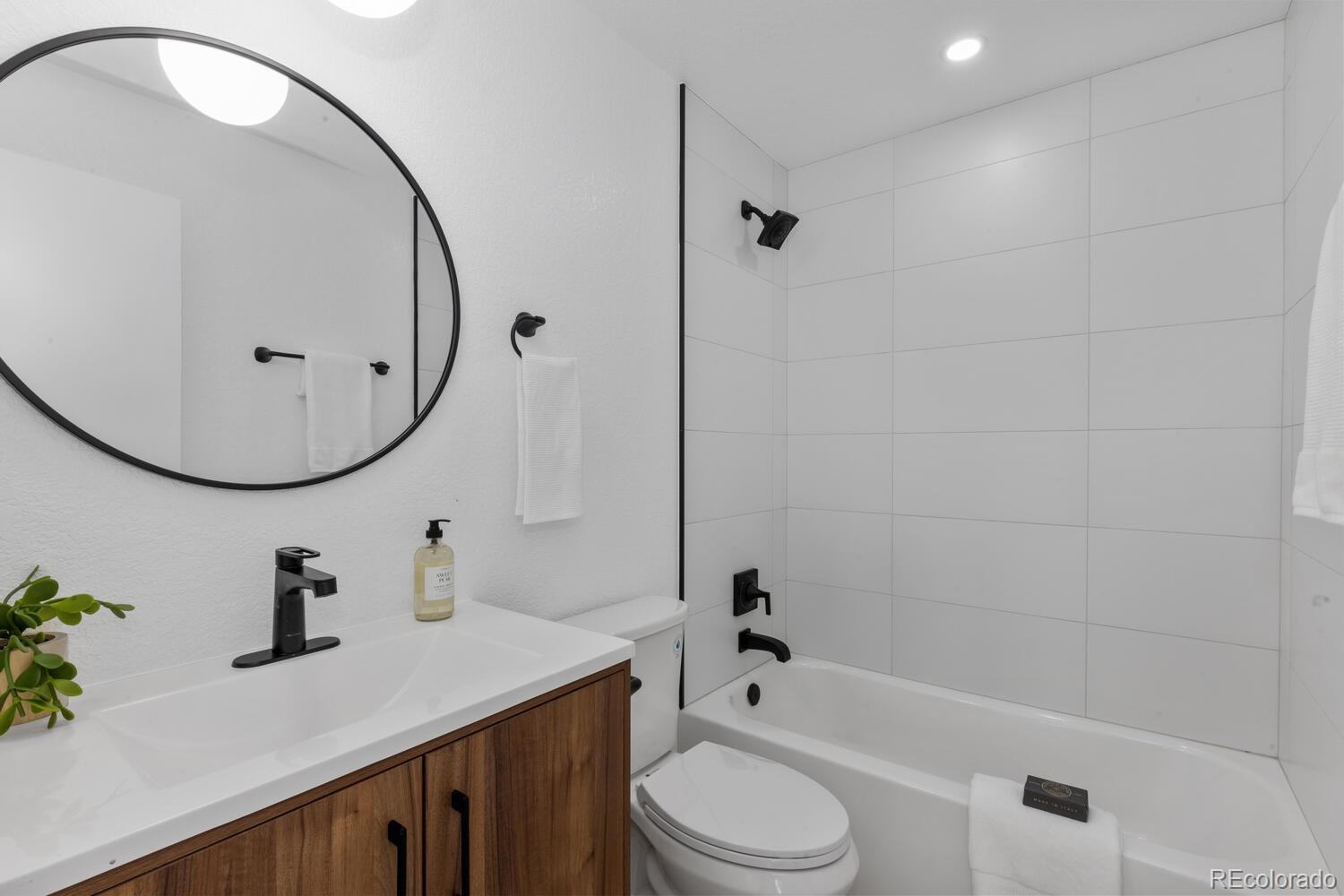 Bathroom with white tiled tub surround, wood vanity, oval mirror, and black fixtures.