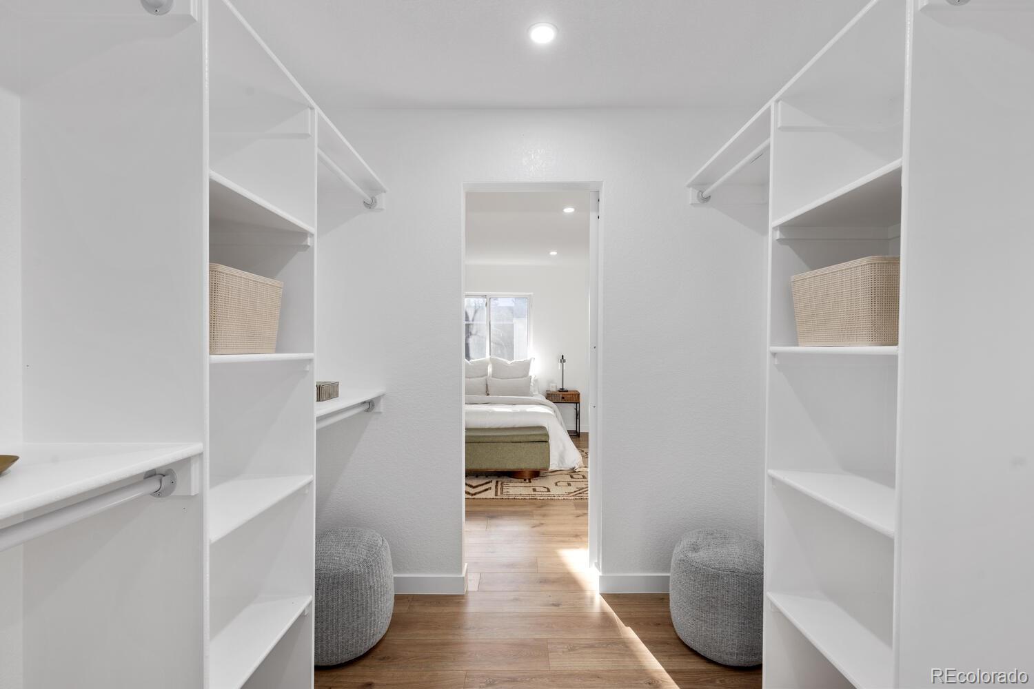 Walk-in closet with white built-in shelving, woven baskets, and gray stools beside a bedroom doorway.