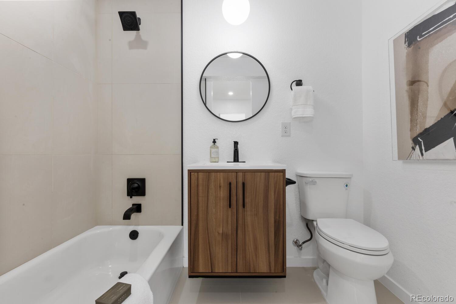 Modern bathroom with a wood vanity, round mirror, tiled bathtub surround, and black fixtures.