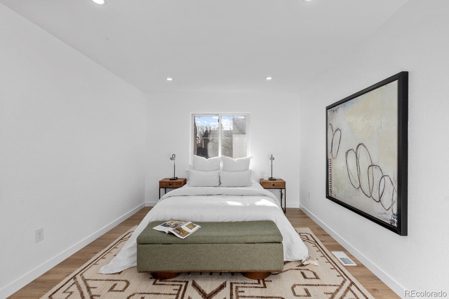 Staged bedroom with a white bed, green bench, patterned rug, and large abstract wall art.