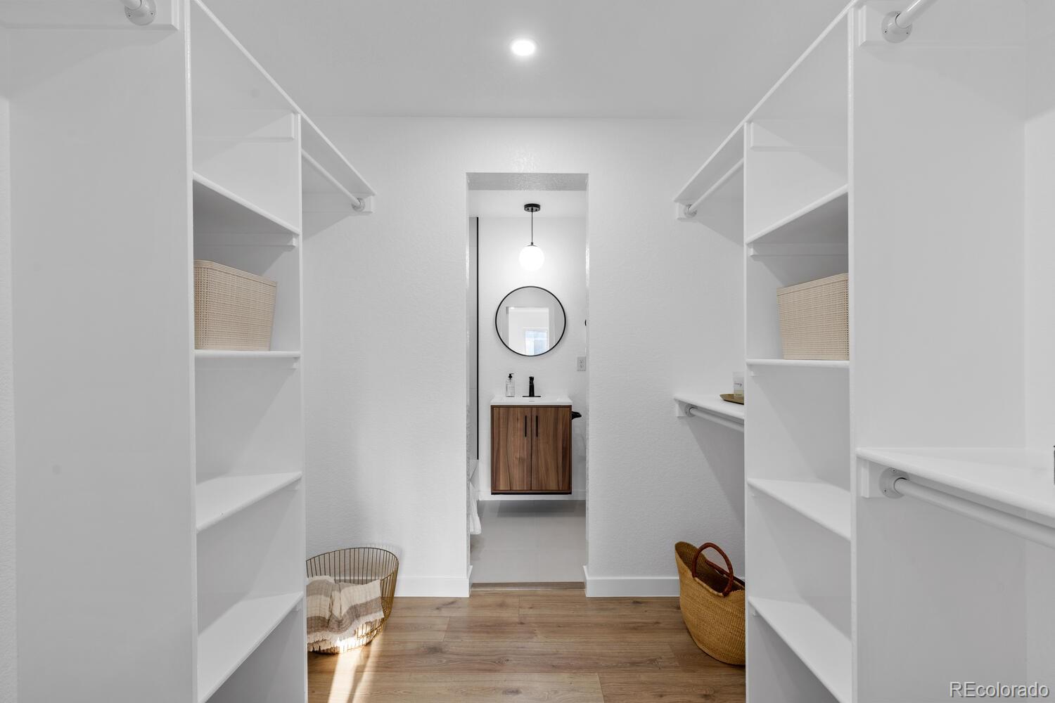 Walk-in closet with white open shelving and wood floors, opening to a bathroom vanity.