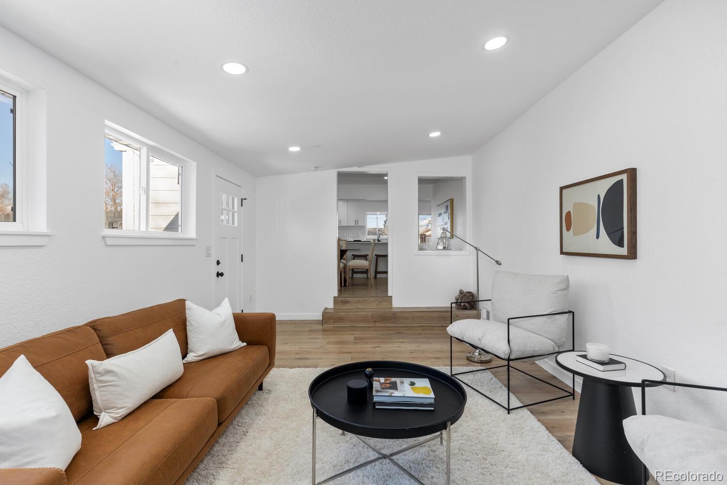 Staged living room with a caramel-brown sofa, white lounge chair, and doorway to the kitchen.