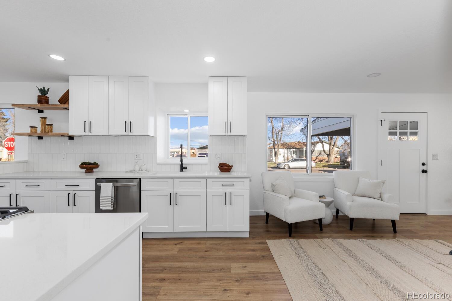 Bright white kitchen and sitting area with wood floors, open shelving, and two armchairs by a window.