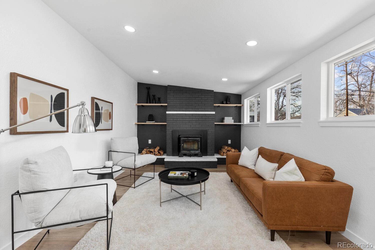 Staged living room with a black brick fireplace, tan sofa, white chairs, and large windows.