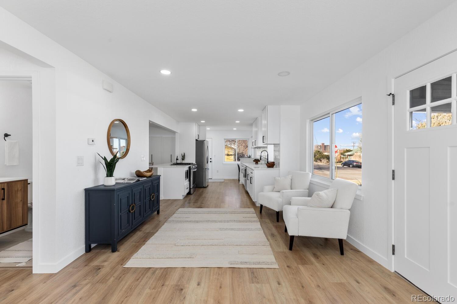 Open-plan kitchen and living area with white cabinetry, light wood floors, and two armchairs beside large windows.