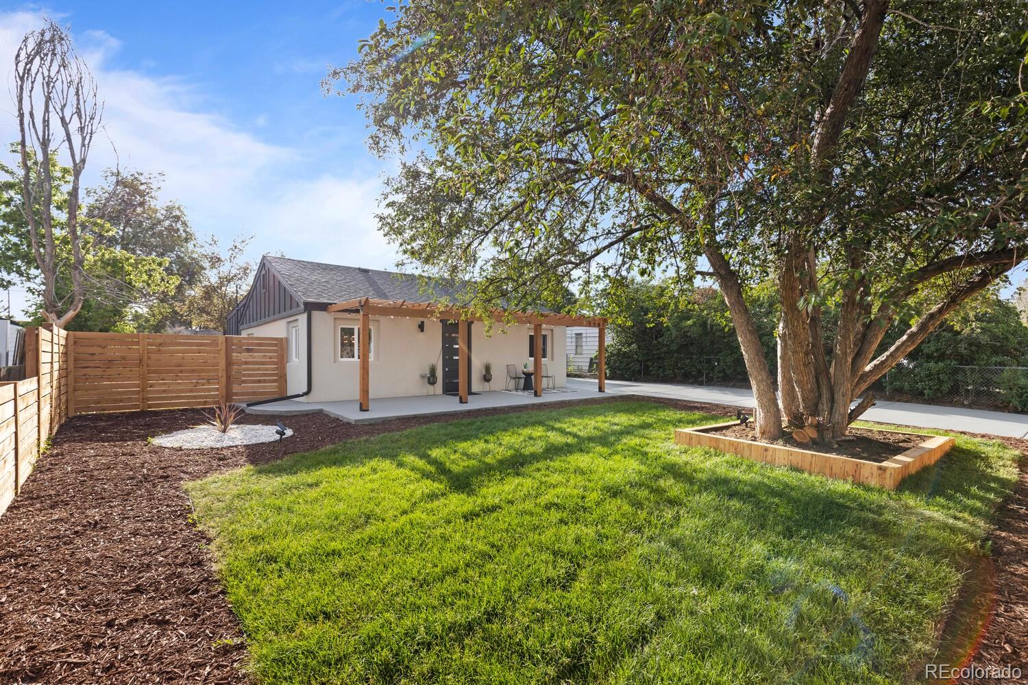 Home’s rear exterior with a pergola-covered patio, fenced lawn, and mature shade trees.