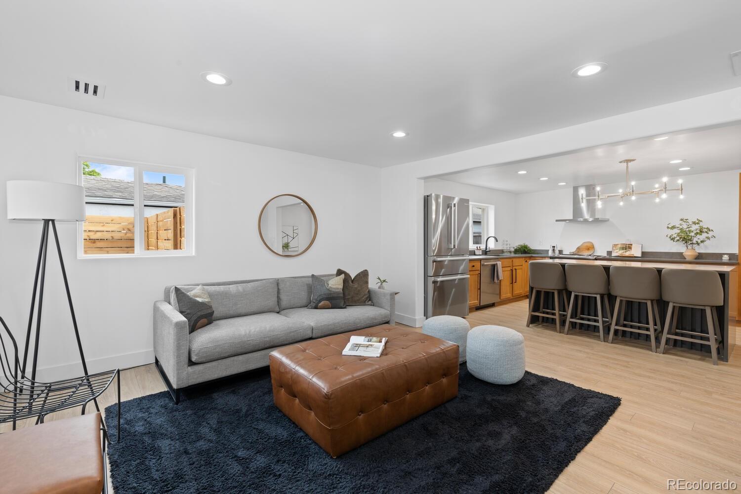 Open-plan living room and kitchen with a gray sofa, leather ottoman, and wood-cabinet island.