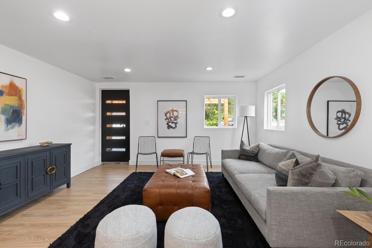 Staged living room with a gray sectional, brown leather ottoman, black rug, and modern black front door.