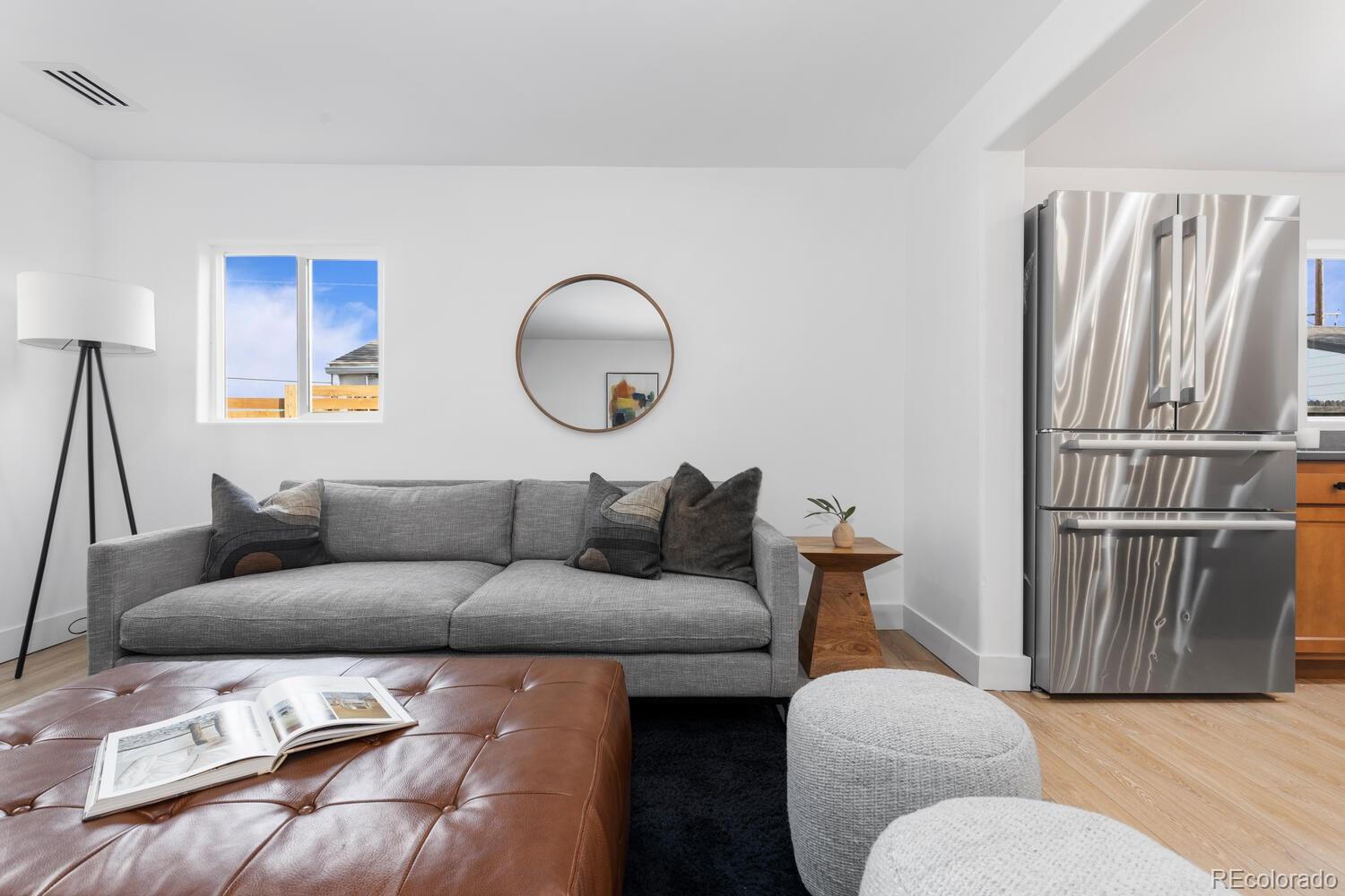 Furnished living room with a gray sofa, round mirror, leather ottoman, and stainless steel refrigerator.