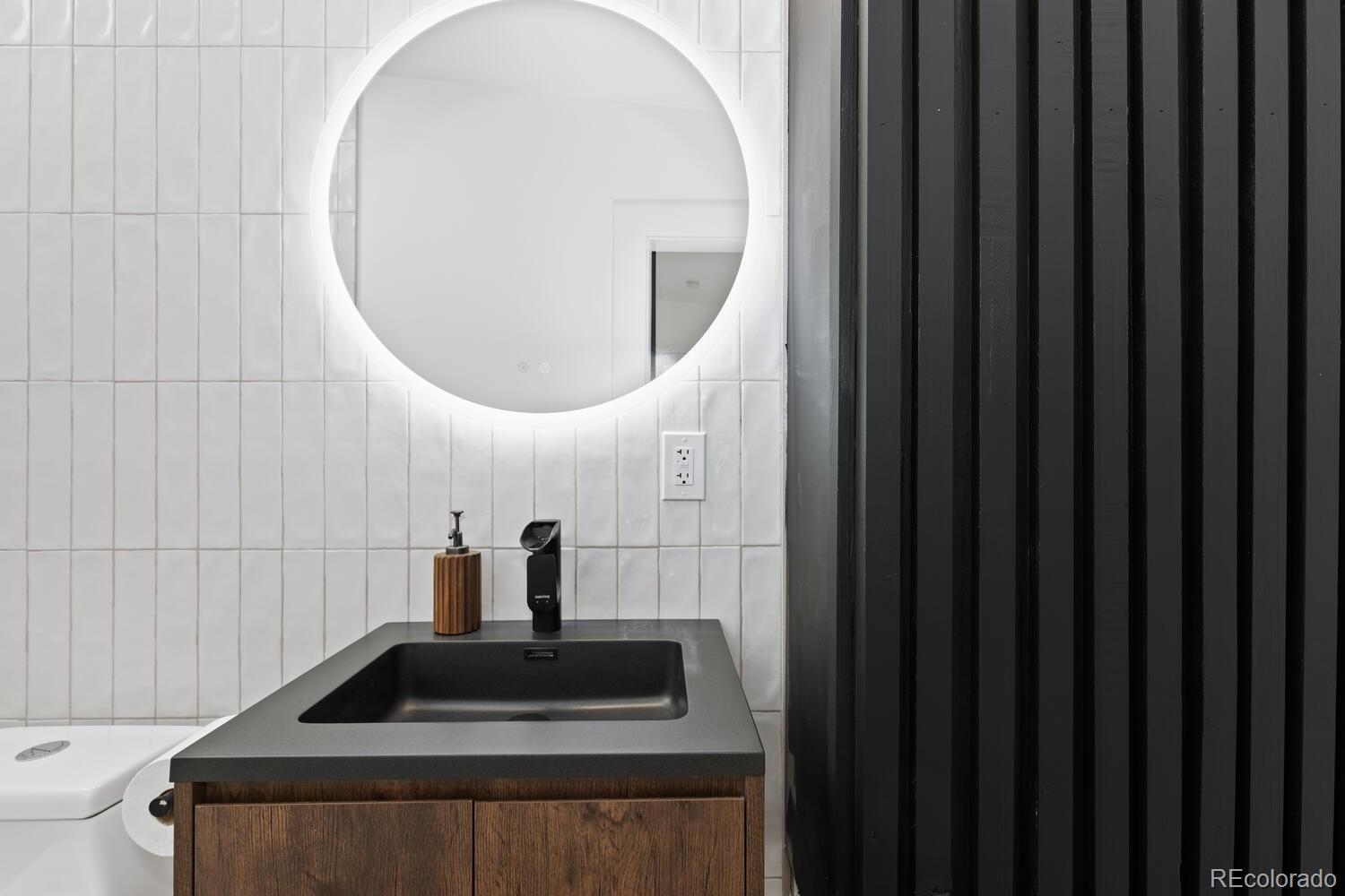 Bathroom vanity with a round illuminated mirror, black sink, wood cabinetry, and black slatted wall.