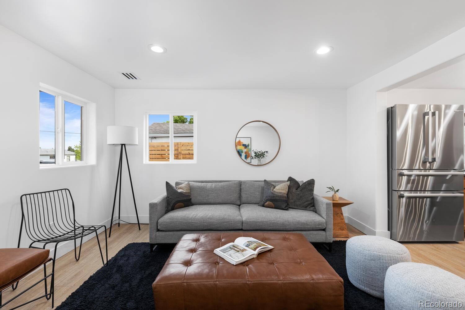 Staged living room with a gray sofa, leather ottoman, large windows, and stainless-steel refrigerator.