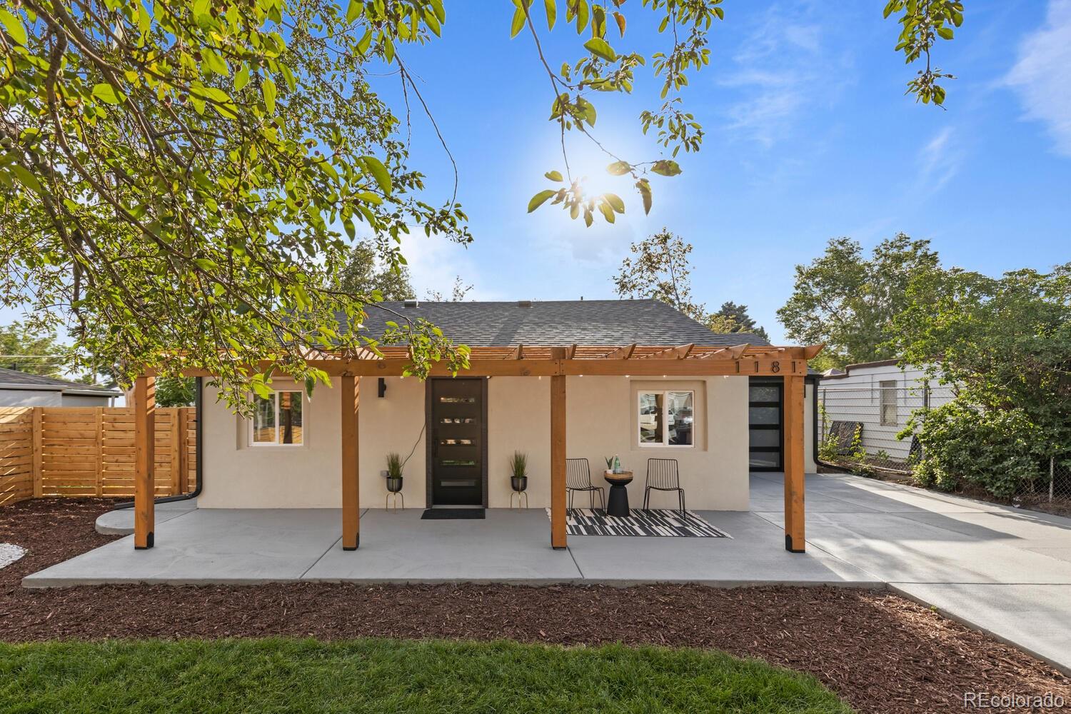 Single-story home's front patio beneath a timber pergola, with outdoor chairs and a wood privacy fence.