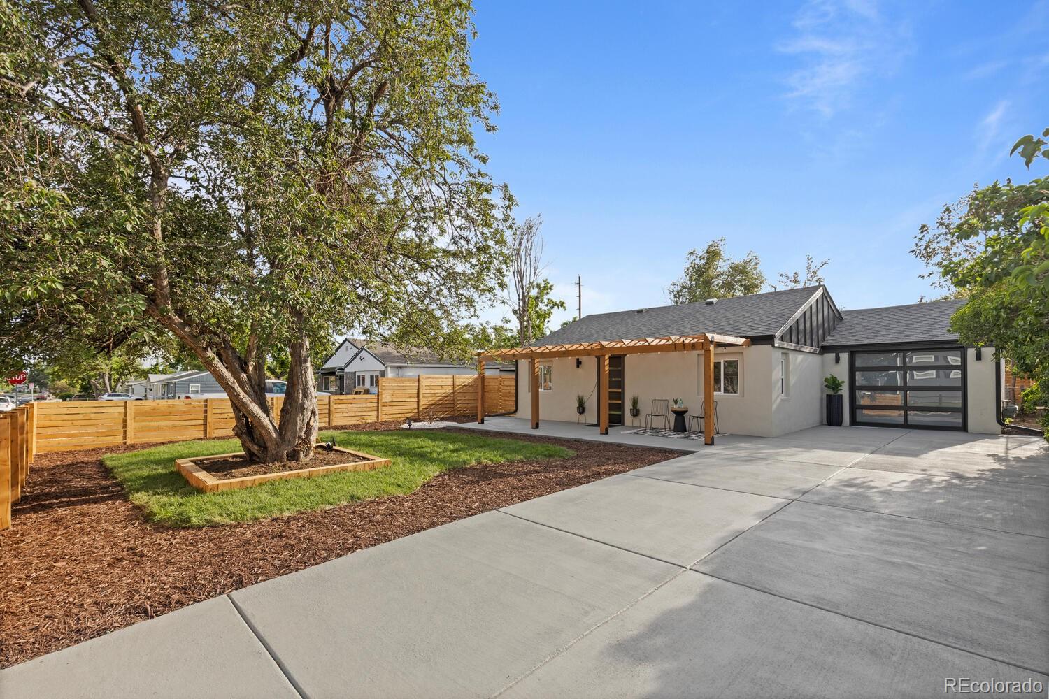 Single-story home with a pergola-covered patio, mature tree, wood fence, and wide concrete driveway.