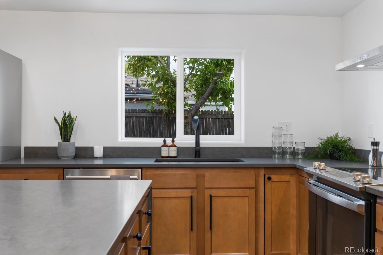 Kitchen with warm wood cabinets, gray countertops, and a sink beneath a window overlooking a leafy tree.