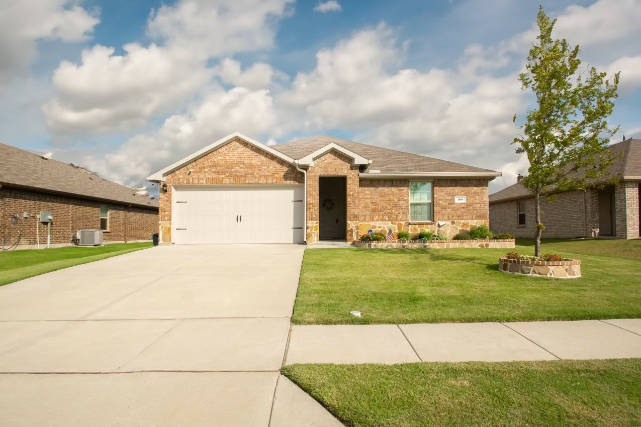 Single-story brick home with a white double garage, broad driveway, and a young tree in the lawn.