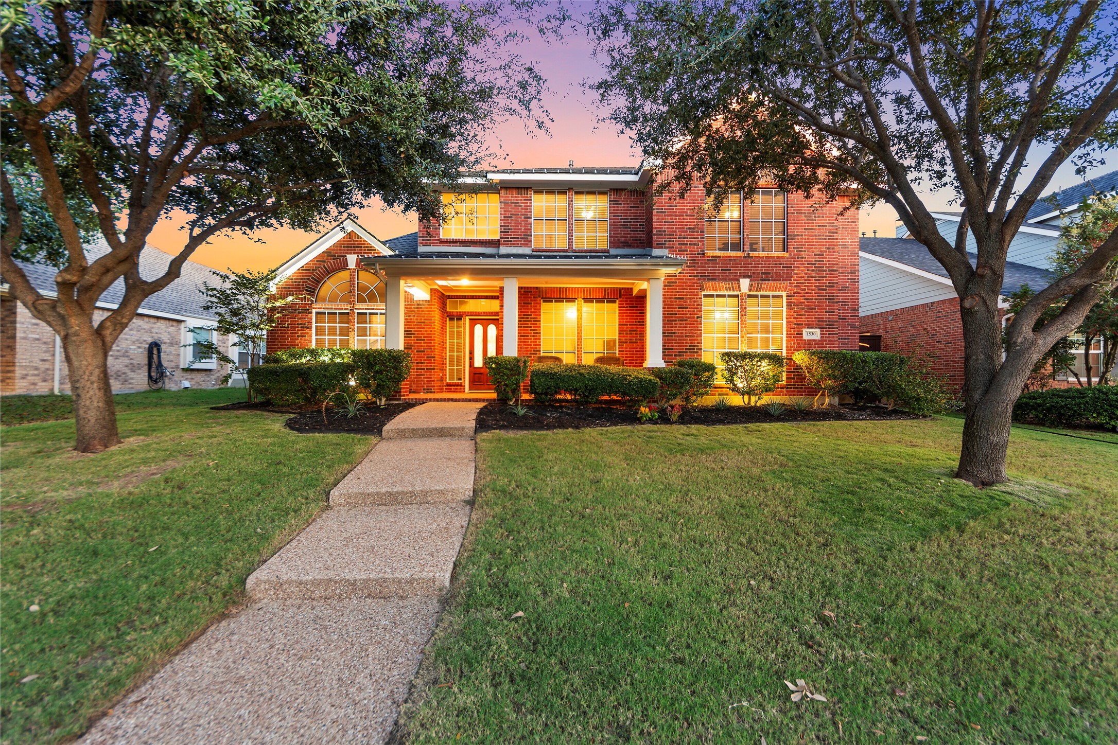 Two-story brick house with warmly lit windows, framed by mature trees at dusk.
