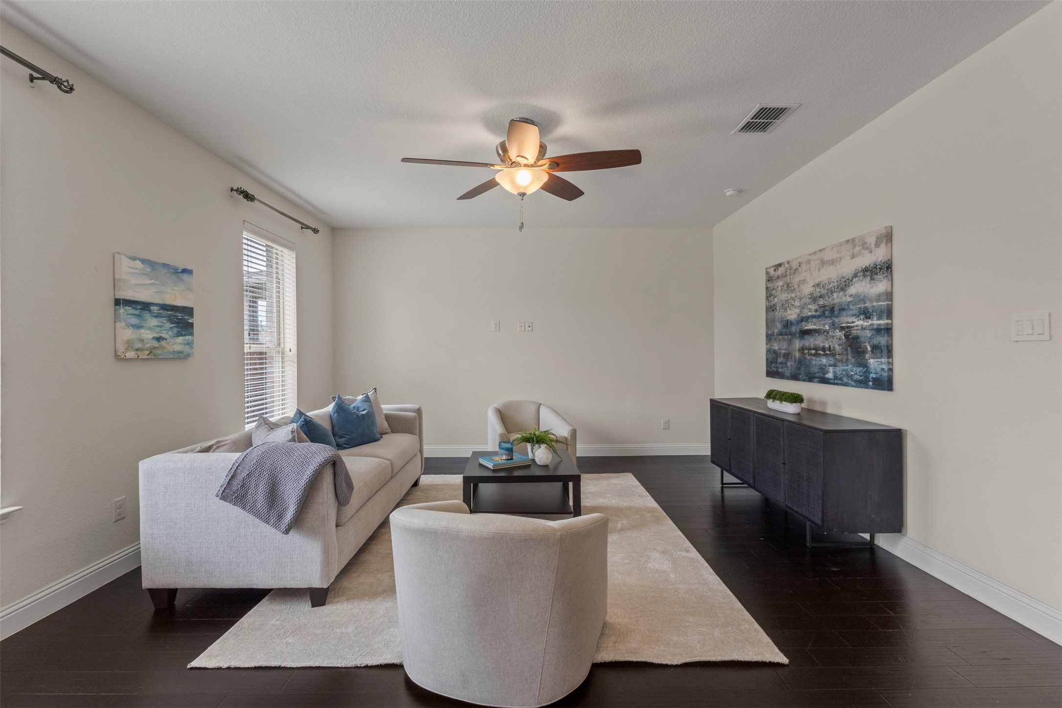 Staged living room with a light sofa, pale rug, dark flooring, and ceiling fan.