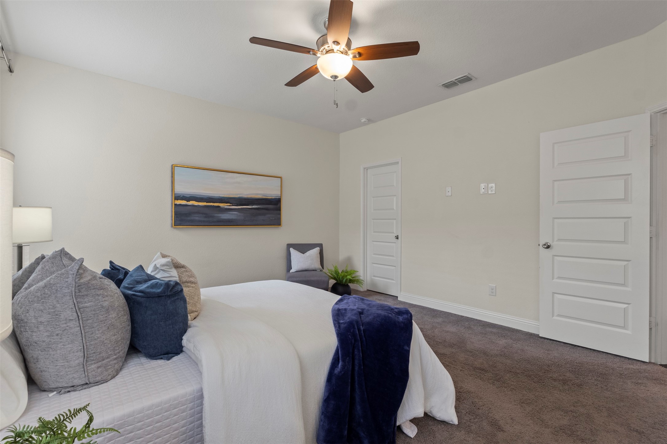 Staged bedroom with a white bed, navy throw, dark-wood ceiling fan, and brown carpet.