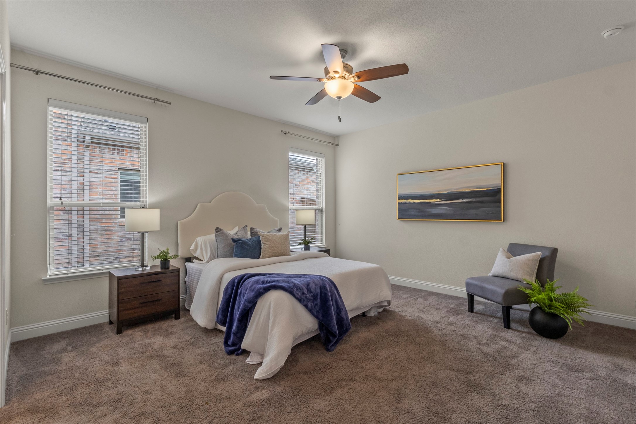 Staged bedroom with a white bed, navy throw, two windows, and gray accent chair.