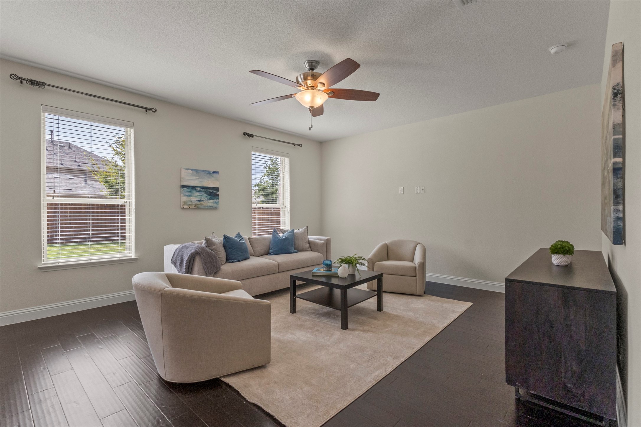 Staged living room with beige seating, dark wood floors, and two bright windows.