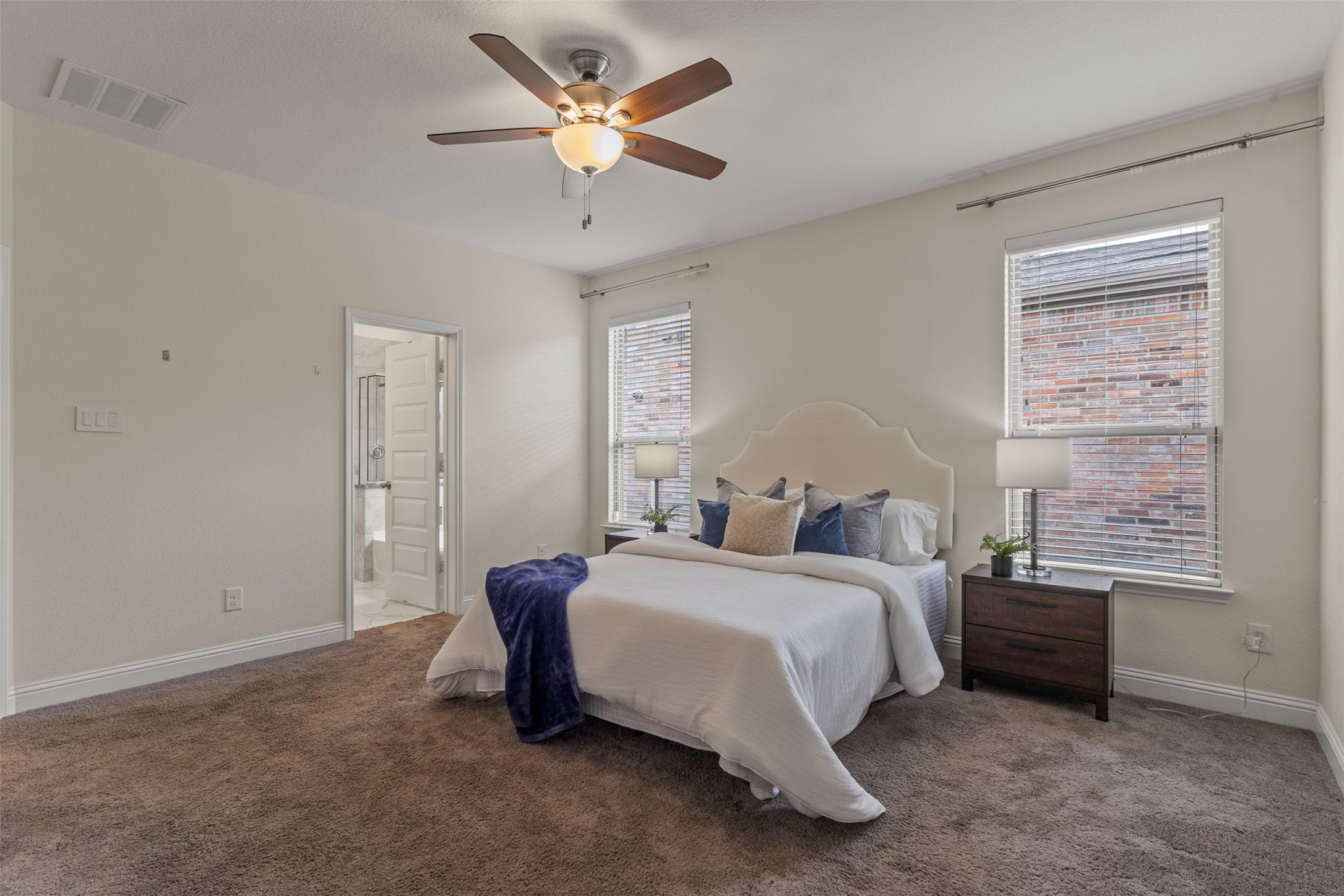 Staged bedroom with a white bed, navy throw, ceiling fan, and windows overlooking a brick wall.