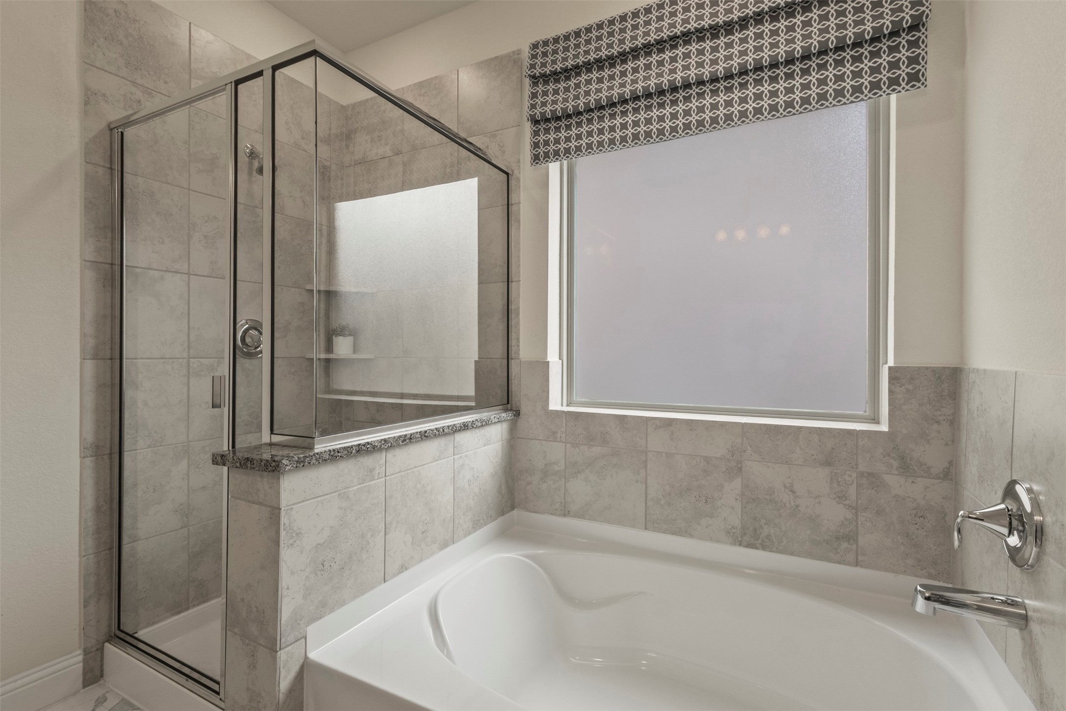 Bathroom with a white corner soaking tub, glass-enclosed shower, gray tile, and a large shaded window.